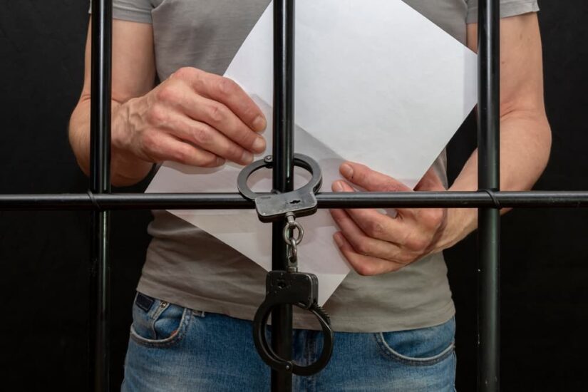 A handcuffed prisoner behind bars holds a white sheet of paper in his hand.