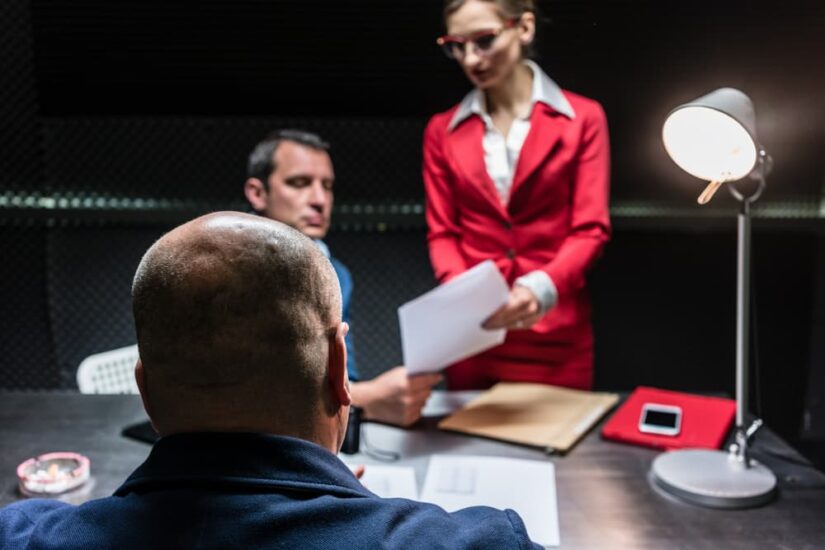 Rear view of a middle-aged suspect or witness sitting at desk during the police interrogation in front of the prosecutor and a female lawyer.